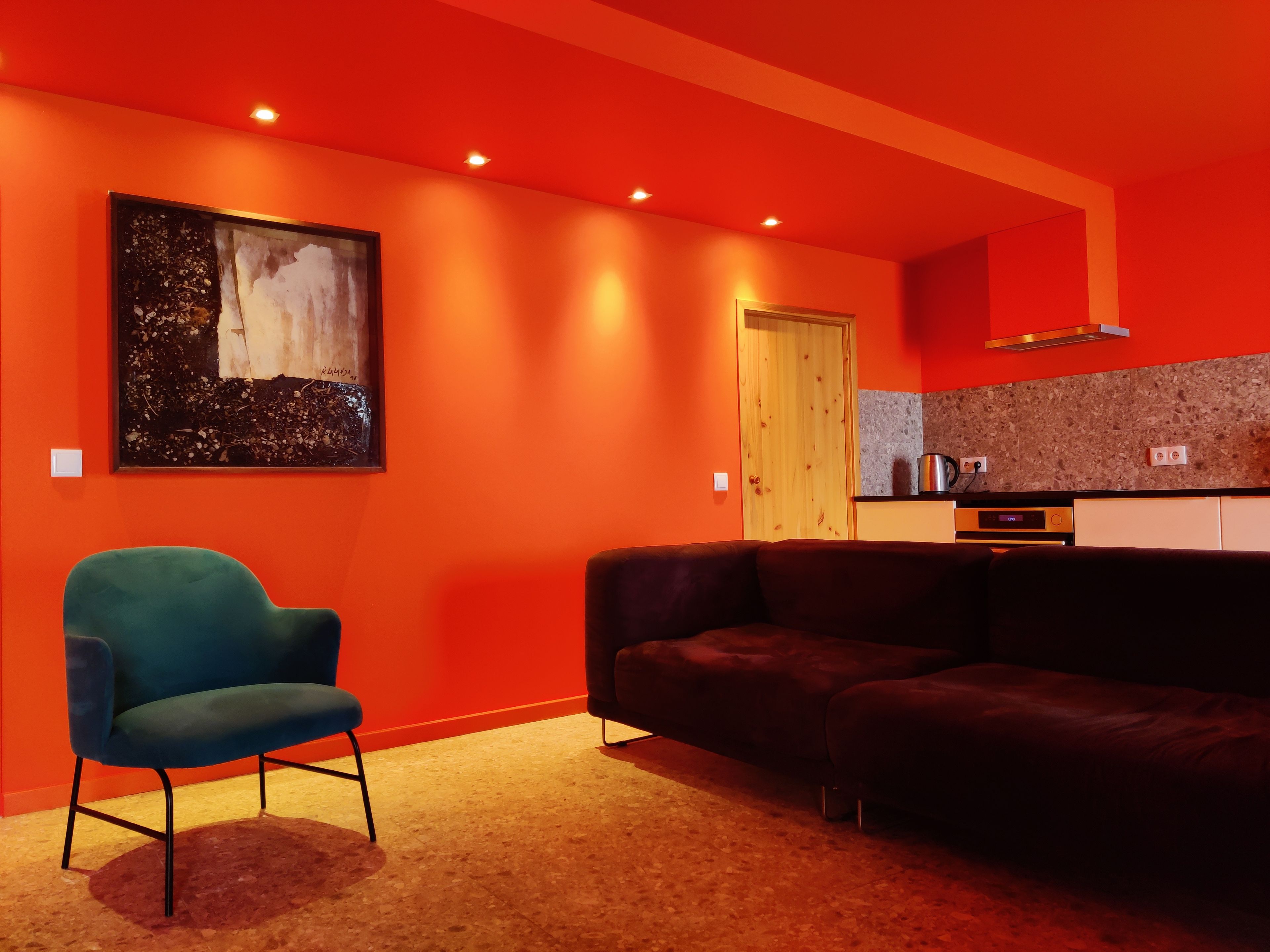 Ein modernes Wohnzimmer mit einer Couch, einem Sessel, einem Gemälde an der Wand, einer Holztür und einem Küchenbereich mit Herd.