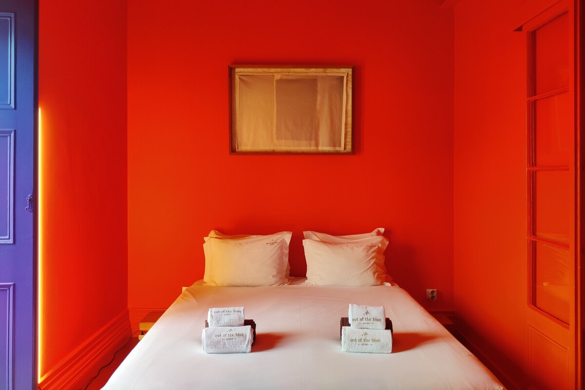 A hotel room with a red wall, a bed with white linens, two towels with text, and a framed picture on the wall.