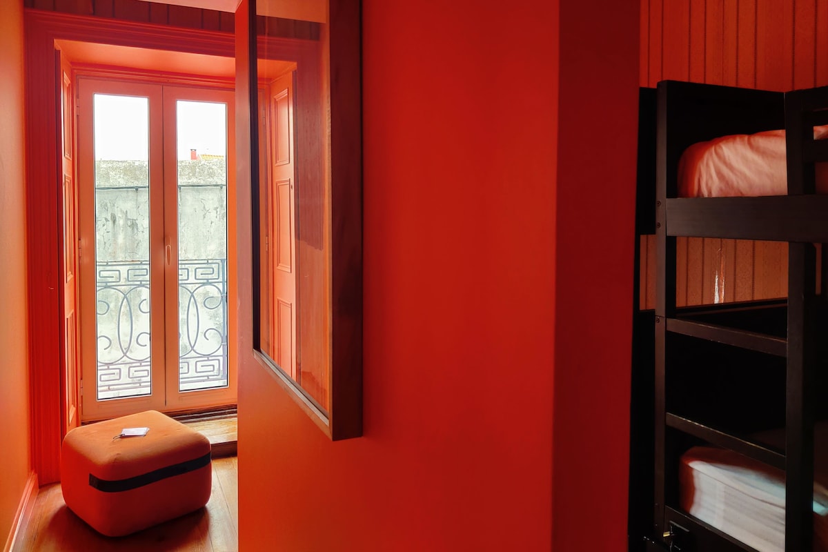 Een kamer met een rode wand, een houten bank, een stapelbed en een deur die leidt naar een balkon met een metalen hekwerk.