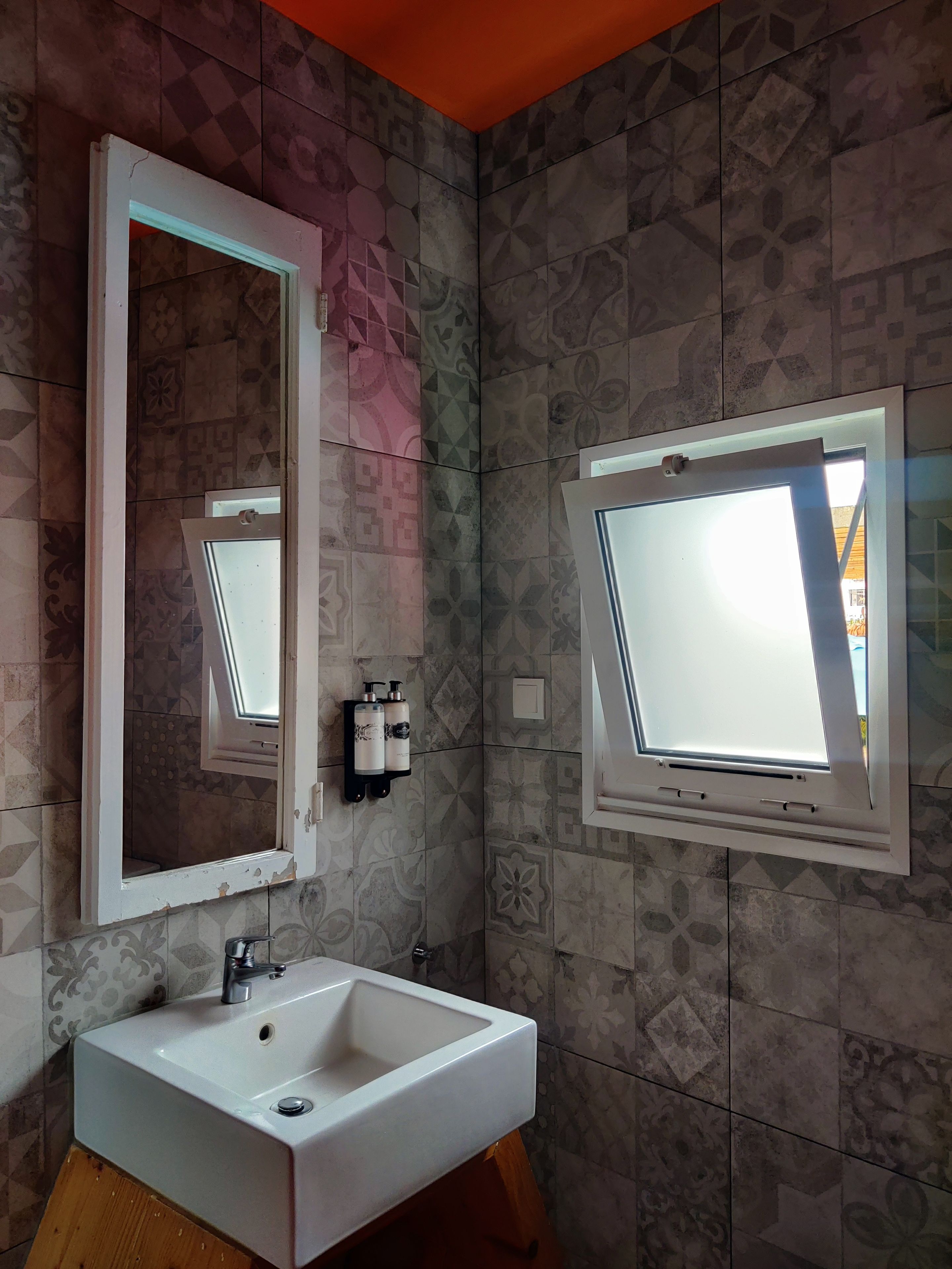 Un baño con un lavamanos blanco, un espejo y dos ventanas.