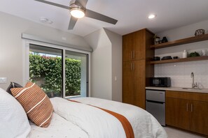 Een slaapkamer met een bed, een plafondventilator, een schuifdeur naar een balkon en een kitchenette met een magnetron en spoelbak.