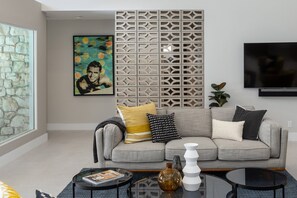 Uma sala de estar moderna com um sofá cinzento, um painel decorativo na parede, uma TV de tela plana e uma mesa de centro de vidro.