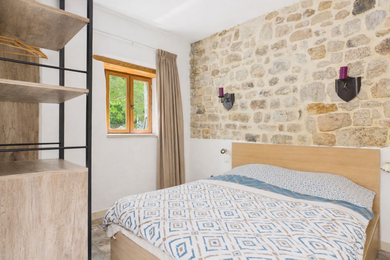 A bedroom with a bed, a wooden shelf unit, a window with curtains, and a stone wall.