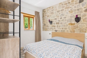 A bedroom with a bed, a wooden shelf unit, a window with curtains, and a stone wall.