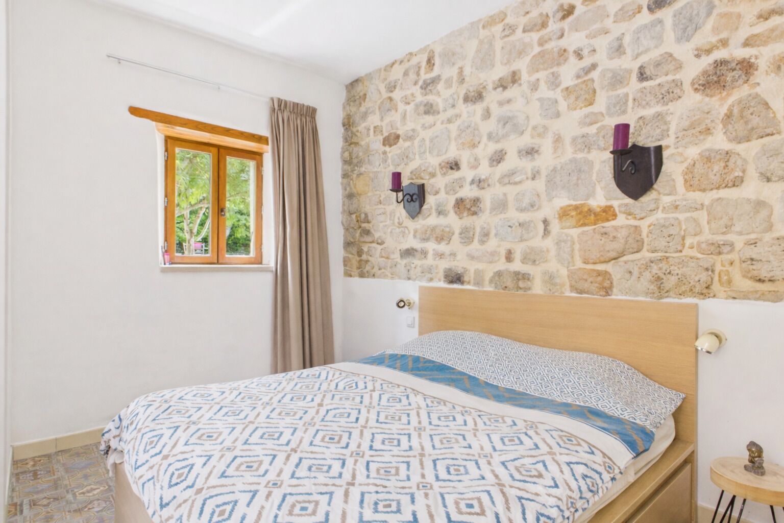 Un dormitorio con pared de piedra, estructura de cama de madera, edredón estampado, ventana con cortinas y una mesita de noche con lámpara.