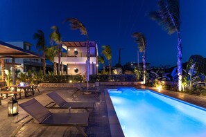 A poolside area with lounge chairs, a swimming pool, and a two-story building with outdoor lighting.