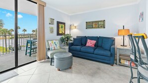 A living room with a blue sofa, a grey ottoman, and a dining table with chairs. There are paintings on the wall and a lamp on a side table. - Gulf Shores Surf & Racquet Club 212A (Gulf Shores)