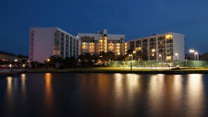 A multi-story hotel with a pool in the foreground, surrounded by a fence and palm trees. - Gulf Shores Surf & Racquet Club 212A (Gulf Shores)
