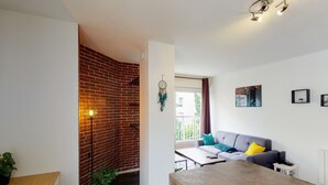 Un espacio habitable moderno con pared de ladrillo, una mesa de comedor de madera, un sofá gris con cojines coloridos y un pequeño balcón visible a través de una ventana.
