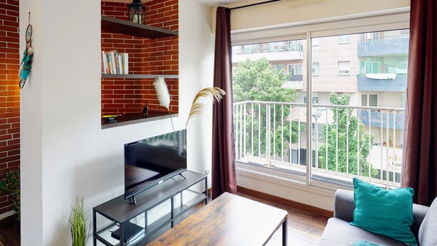 Una sala de estar moderna con un televisor de pantalla plana sobre un mueble negro, una estantería llena de libros, una planta en maceta y vistas a un balcón con barandilla y vegetación al exterior.