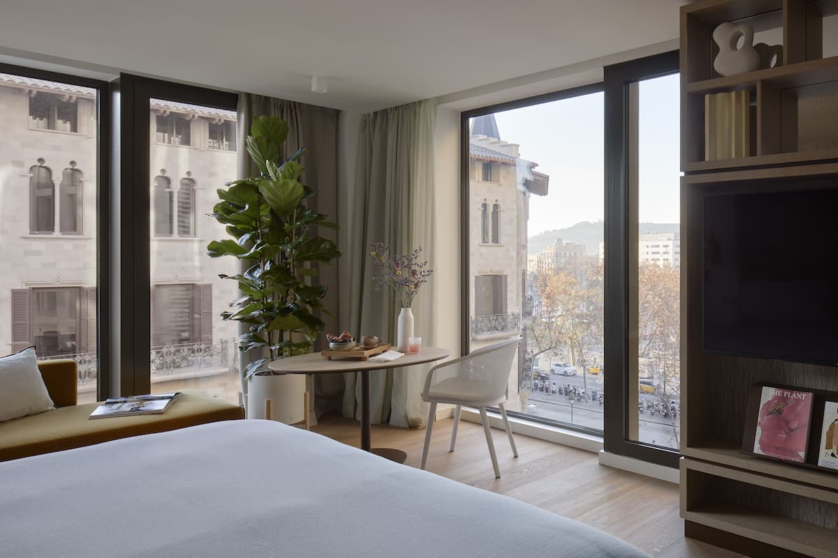 A hotel room with a large bed, a dining table, a chair, a bookshelf, and a large window offering a city view.