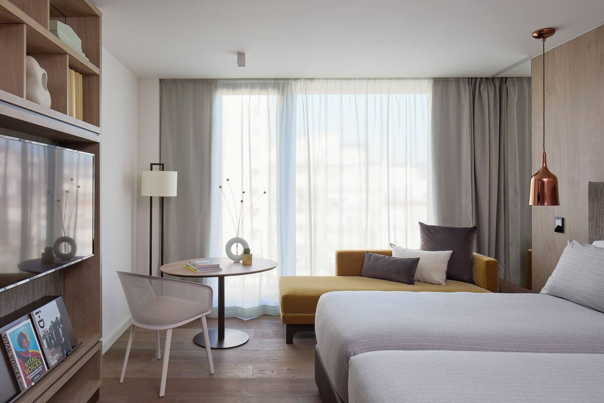 A hotel room with a bed, a sofa, a desk with a chair, a bookshelf, and a large window with curtains.