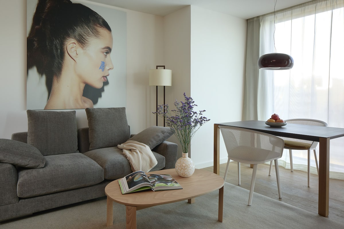 A modern living room with a grey sofa, a coffee table with a magazine, a vase with flowers, and a dining area with a table and chairs.