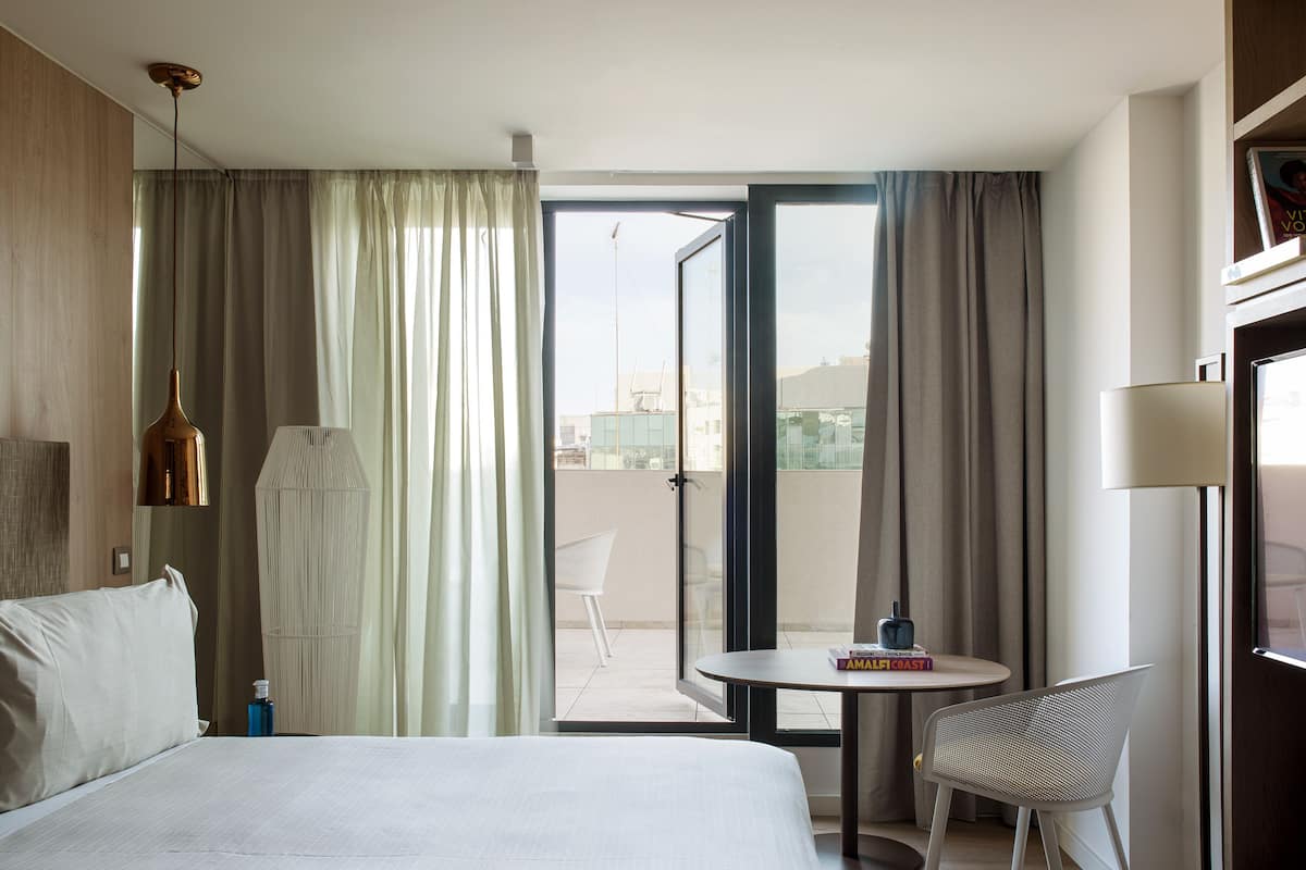 A hotel room with a bed, a chair, a table, and a view through a sliding door.