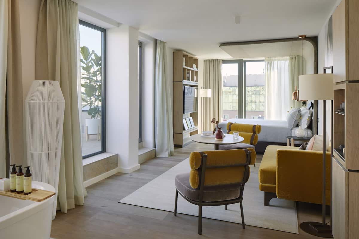 A modern hotel room with a large bed, a sofa, a dining table, and a small kitchen area.