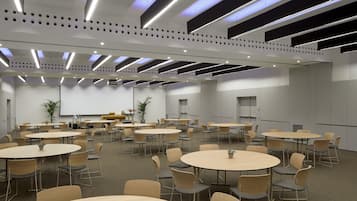 Un salón de conferencias con varias mesas redondas y sillas, equipado con una pantalla de proyector y zona de presentaciones.