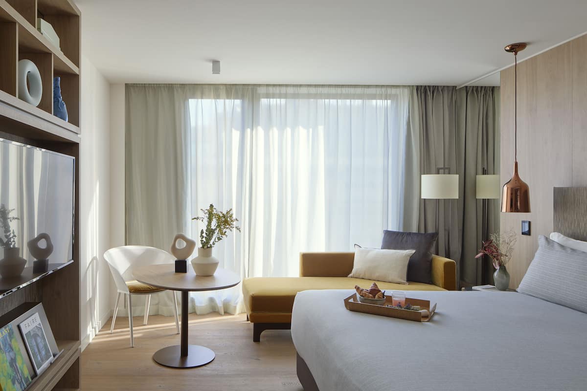 A hotel room with a bed, a round table with a chair, a small sofa, a potted plant, and a tray with pastries.