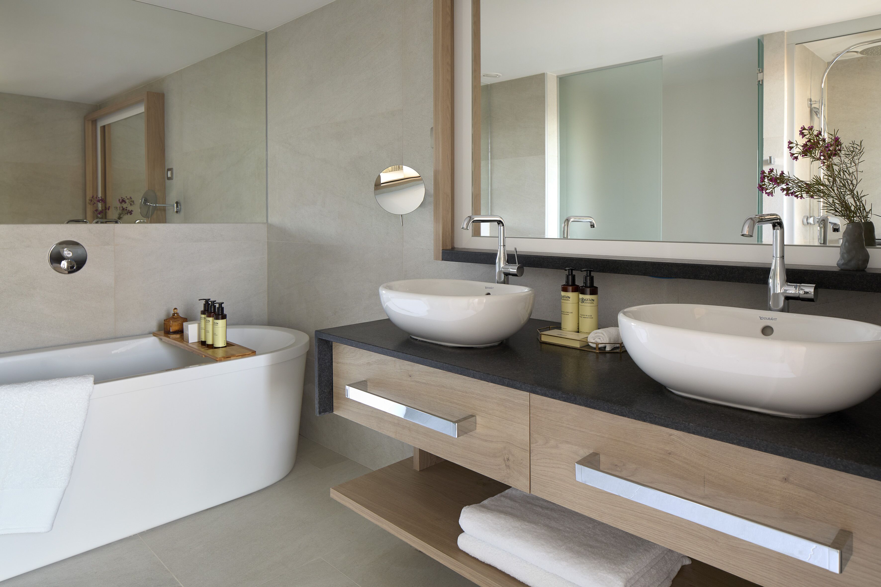 Un baño moderno con dos lavamanos blancos, una bañera grande y un mueble de tocador de madera con cajones.