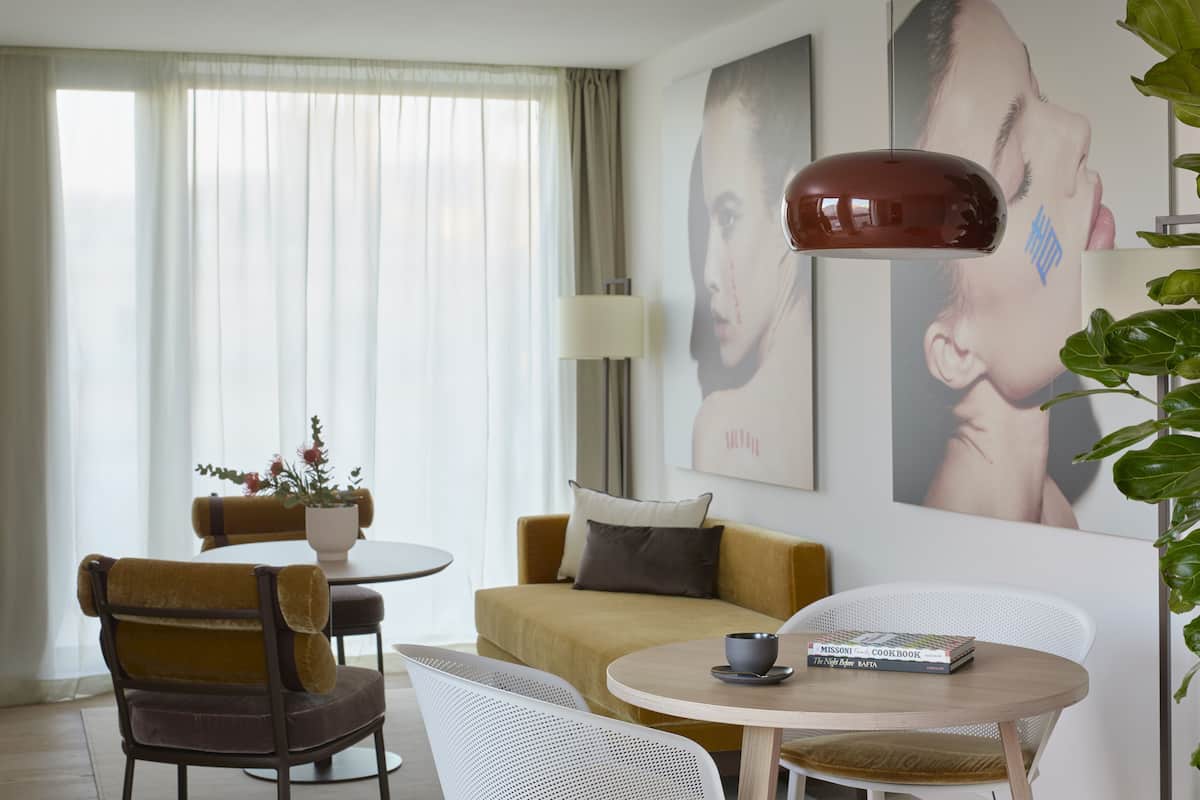 A modern hotel room with a round dining table, a sofa, and a large window with sheer curtains.
