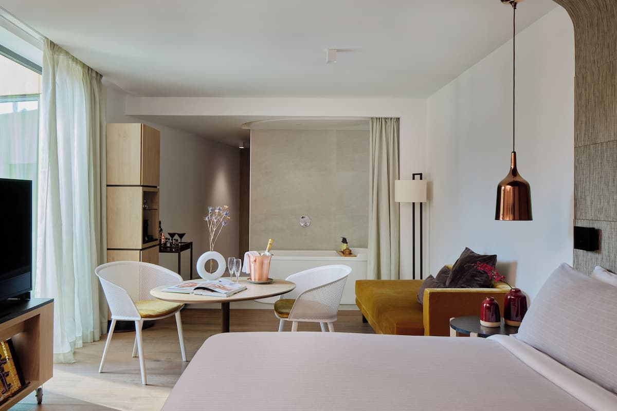 A modern hotel room with a bed, two white chairs, a small round table with a magazine, a yellow sofa, and a large window with curtains.