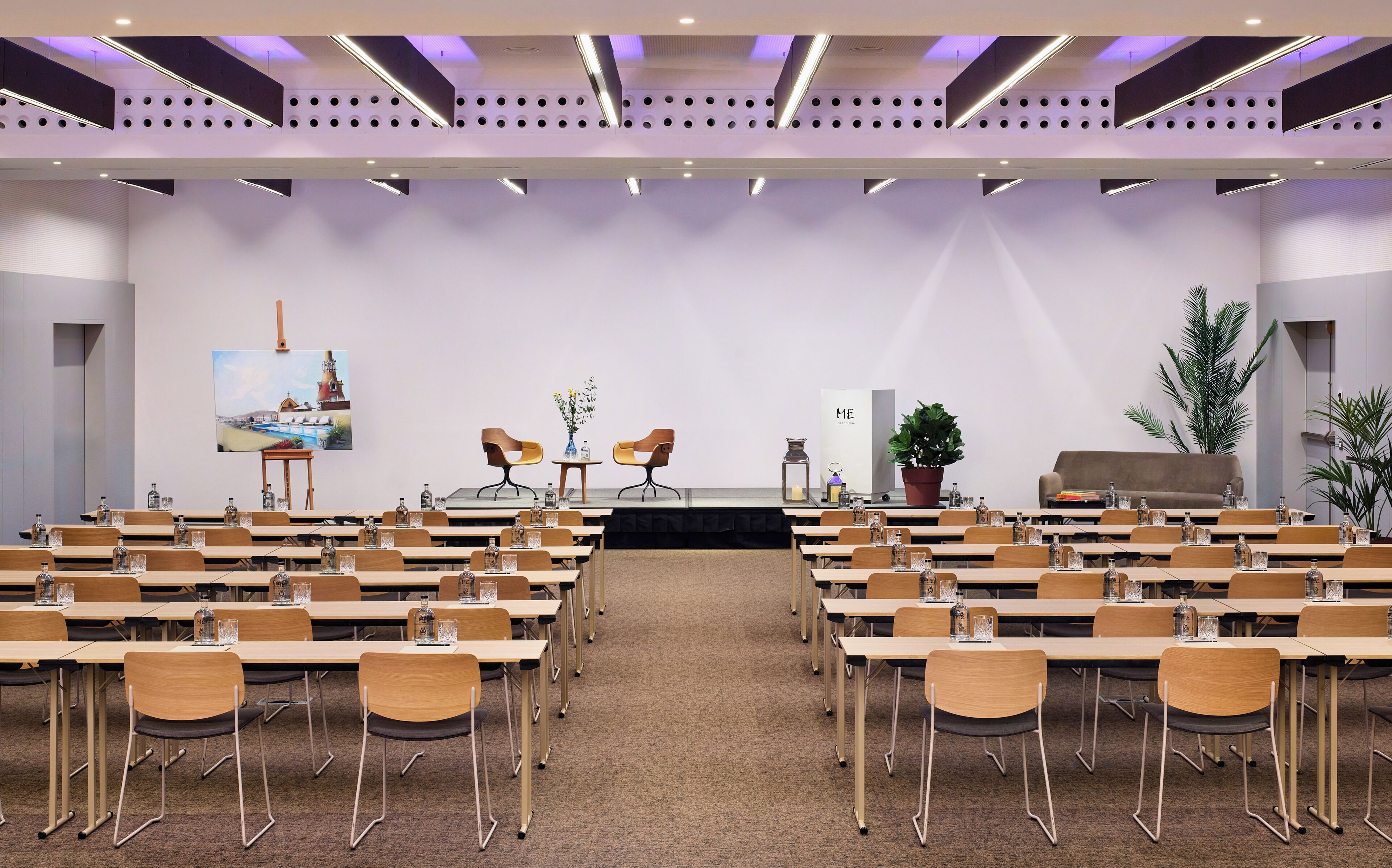 Un salón de conferencias con varias mesas y sillas, un escenario con dos sillas y un cuadro grande en la pared.