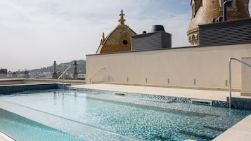 A swimming pool with clear water, surrounded by a low wall and a beige building with a tiled roof.