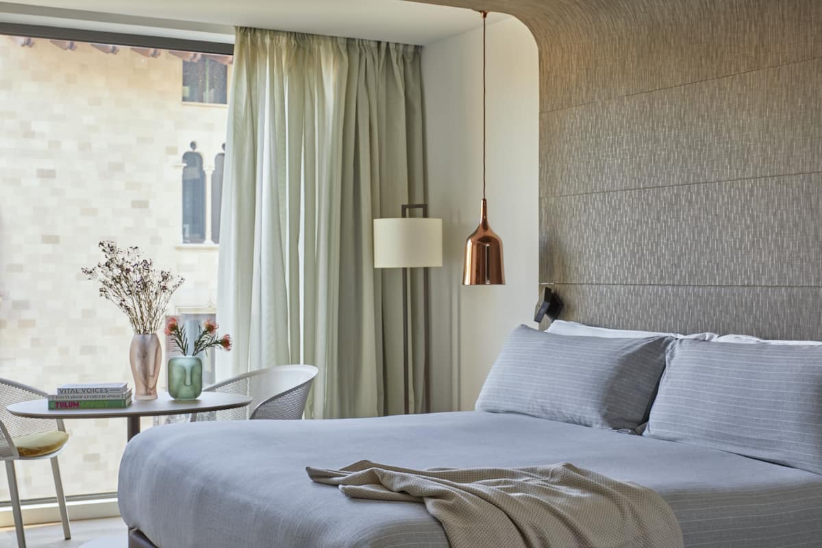 A modern hotel room with a bed, a table with books, a vase with flowers, and a view of a building through the window.
