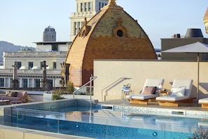 Zona de piscina en la azotea con sillones de descanso, una mesita y vistas a un edificio histórico al fondo.