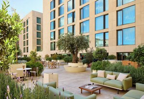 A modern outdoor lounge area with seating, a central tree, and a building in the background. - ME Barcelona (Barcelona)