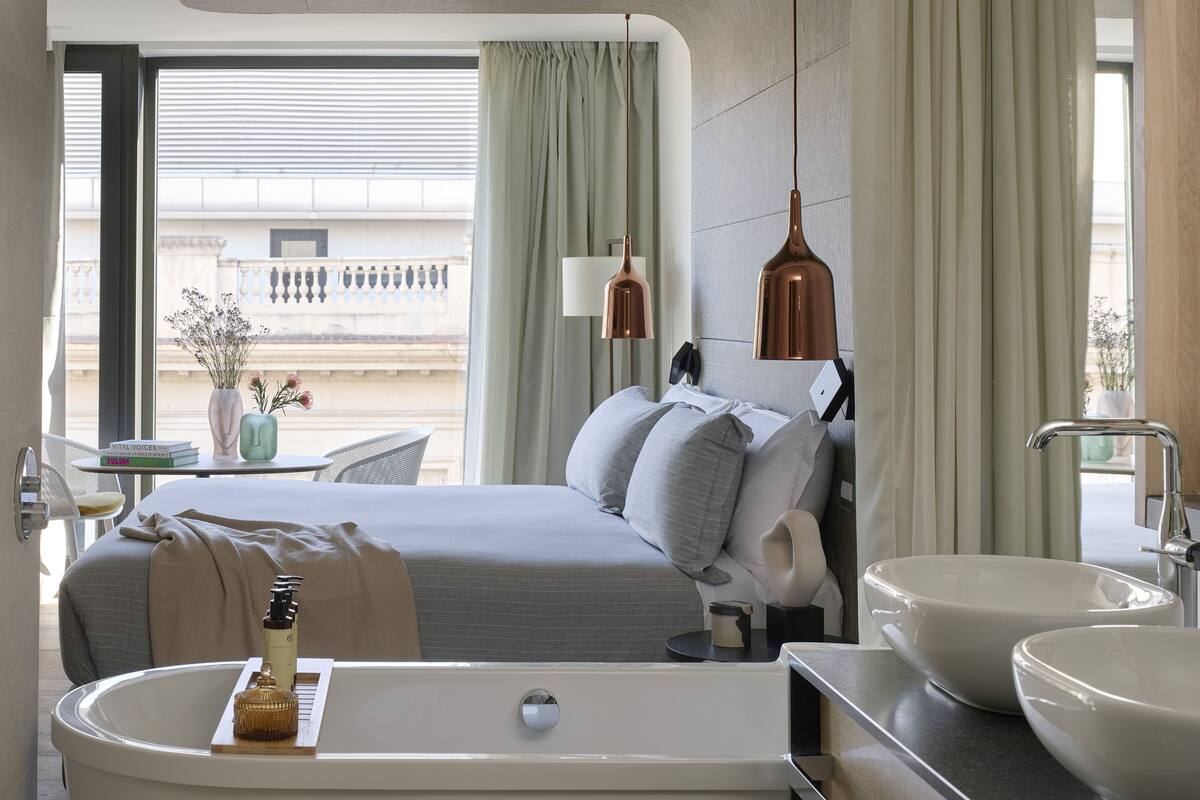 A modern hotel room with a large bed, a bathroom with a bathtub and two sinks, a bedside table with a lamp, and a view of the cityscape.