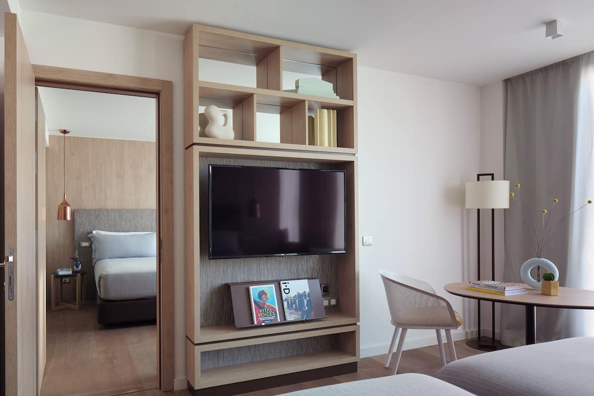 A modern hotel room with a large flat-screen TV, a wooden shelving unit with books and decorative items, a comfortable bed, a small bedside table with a lamp, and a view of the city through the window.