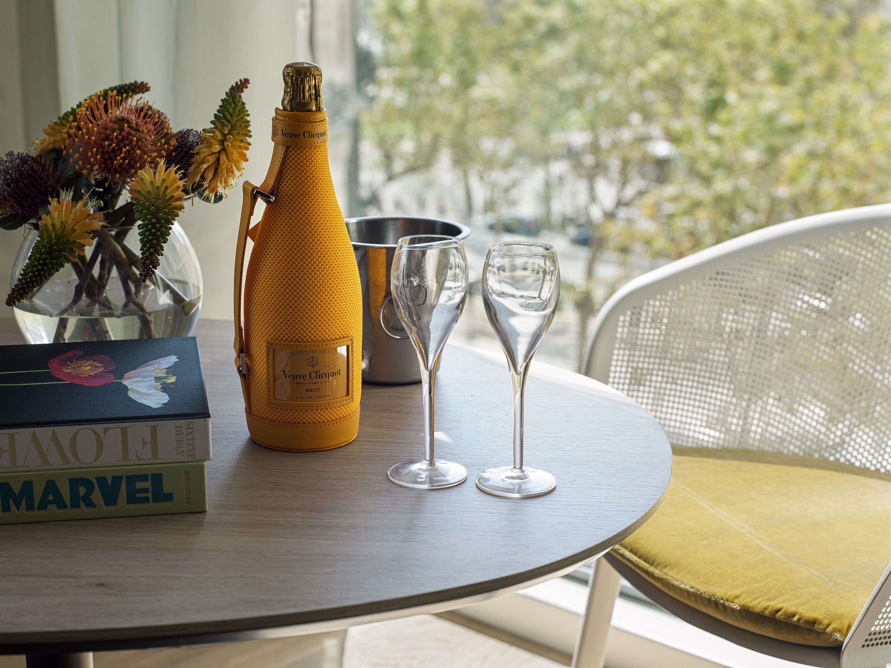 A table with a bottle of Veuve Clicquot champagne, two wine glasses, a metal bucket, a vase with flowers, and a Marvel book.