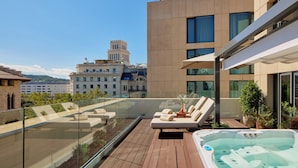Une terrasse sur le toit dotée d’un jacuzzi, de chaises longues et d’une balustrade en verre offrant une vue sur le paysage urbain.