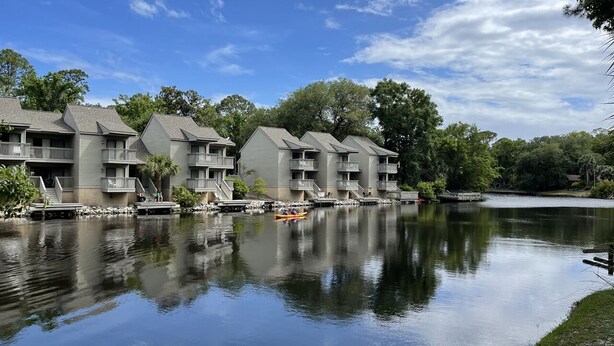 Una hilera de edificios de varios pisos con balcones, un lago tranquilo y un muelle con un bote pequeño.