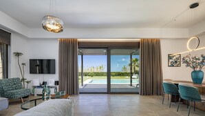 Un soggiorno moderno con una grande TV, un divano, un'area pranzo con sedie e vista su una piscina attraverso ampie vetrate.