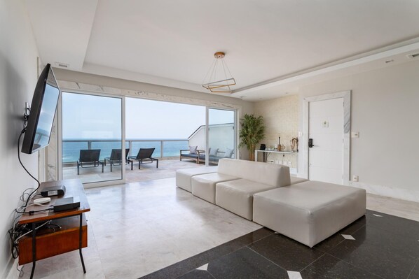 A modern living room with a large window overlooking the sea, a white sofa, a desk with a computer, and a TV mounted on the wall. - Penthouse Overlooking Ipanema Beach Vsc2 (Rio de Janeiro)