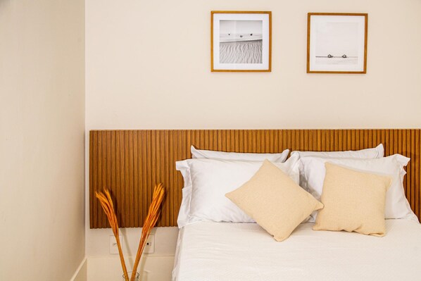 A neatly made bed with white linens and two beige pillows, a wooden headboard, and two framed pictures on the wall.