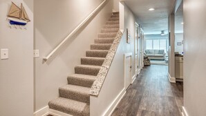 Un escalier recouvert de moquette, doté d’une rampe, mène à un couloir au plancher de bois offrant une vue sur un salon avec un canapé et une fenêtre.