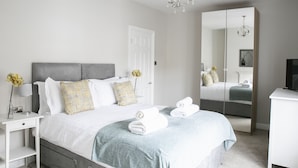 A modern bedroom with a large bed, bedside tables, a dresser, and a mirror. - Deluxe 4 bedroom house (Harrogate)