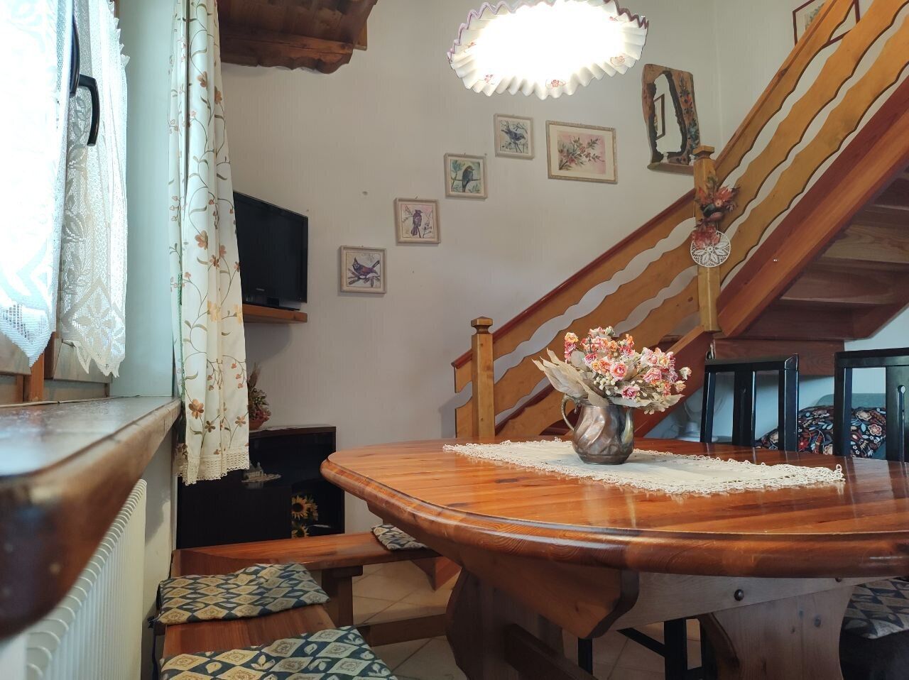 A wooden dining table with a vase of flowers, a lace table runner, and a wooden staircase with a decorative railing.