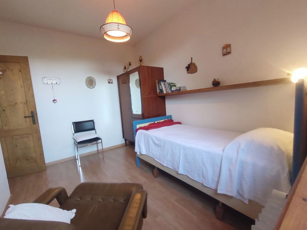 Quarto pequeno e simples com uma cama de solteiro, uma cadeira, um guarda-roupa de madeira e um candeeiro suspenso.