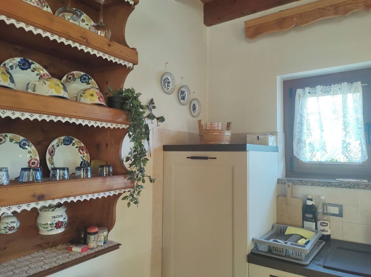 Eine Küche mit Holzregalen, die Teller und Gläser beherbergen, einem Kühlschrank und einem Fenster mit Spitzenvorhängen.