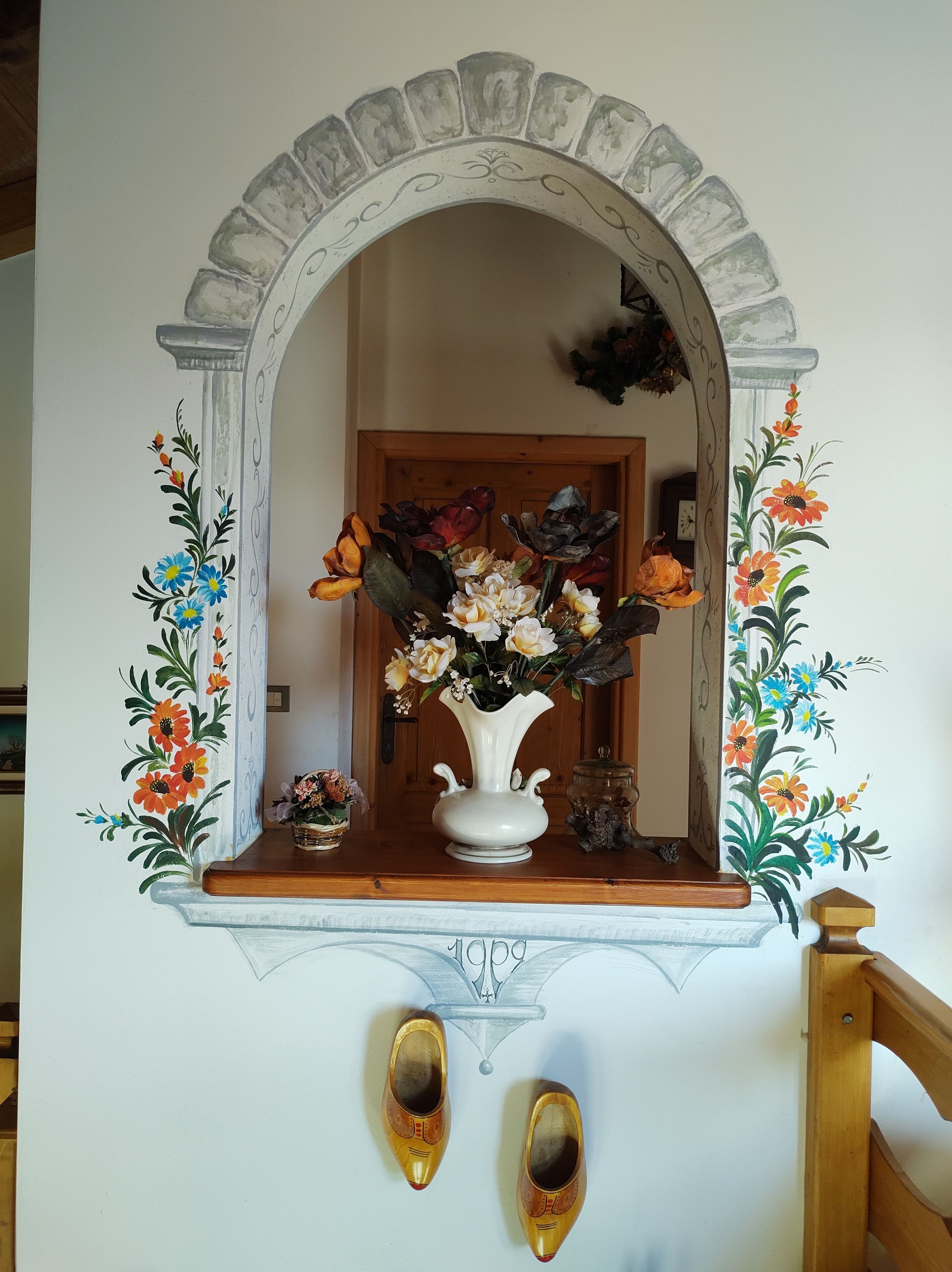 Un miroir orné d’un arc décoratif, un vase rempli de fleurs et une paire de chaussures placée en dessous.