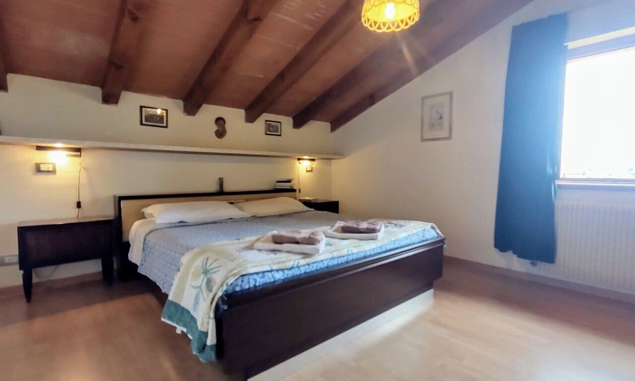 Une chambre avec un plafond en bois, un lit recouvert d’une courtepointe bleue et blanche, deux tables de chevet avec des lampes et une fenêtre ornée de rideaux bleus.