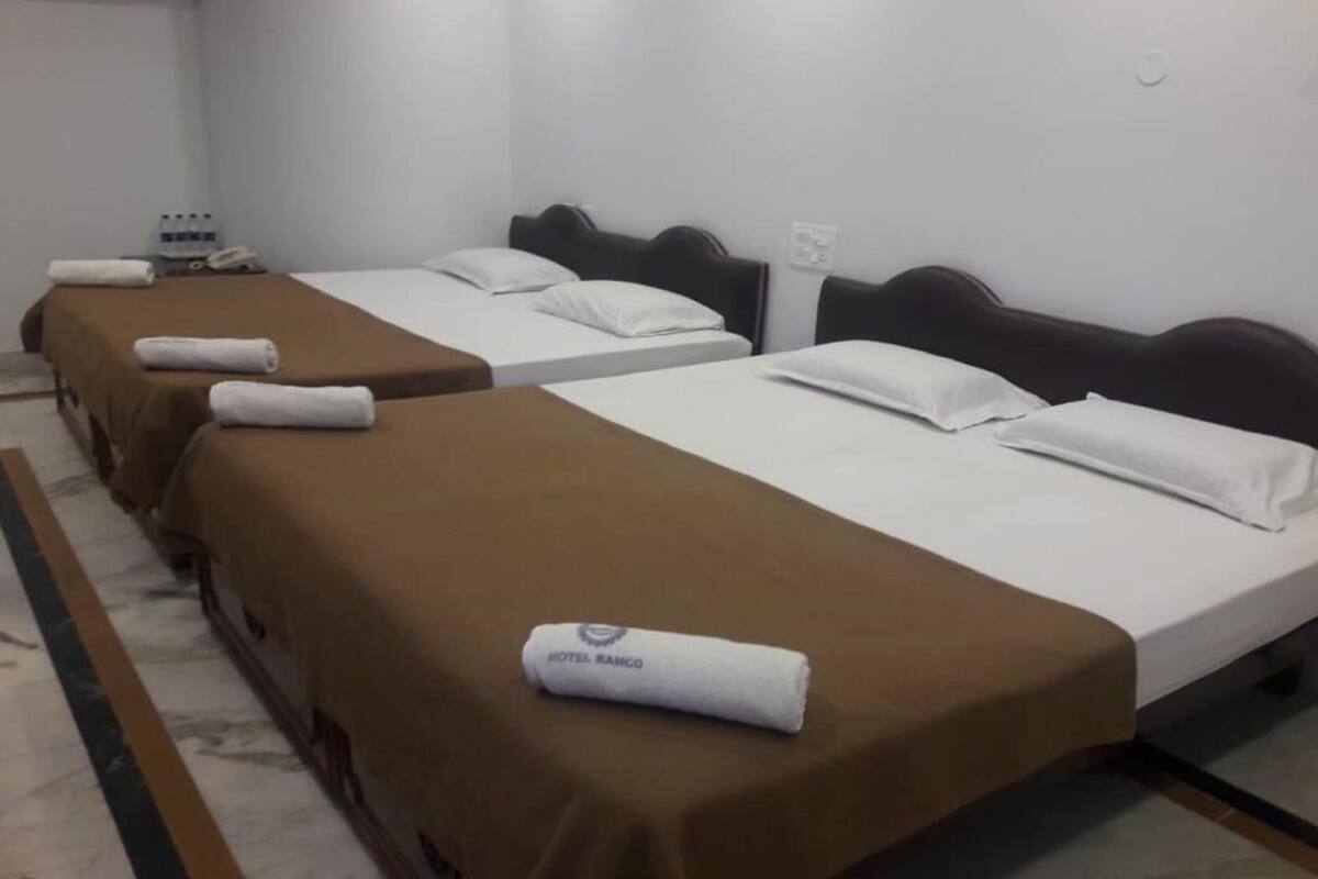 A hotel room with three beds, each with a brown bedspread and white pillows. A rolled towel with "HOTEL RAMCO" is placed on the first bed.