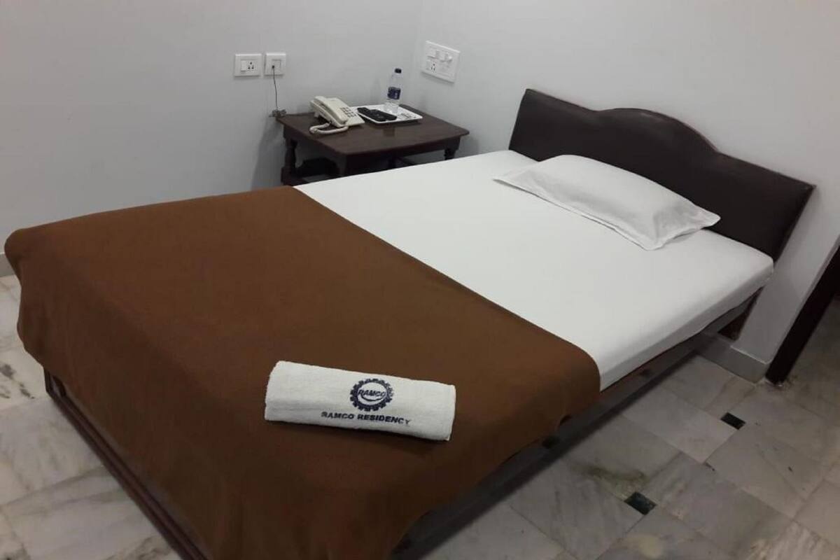 A hotel room with a bed, a nightstand with a phone and a bottle, and a brown blanket with a logo.