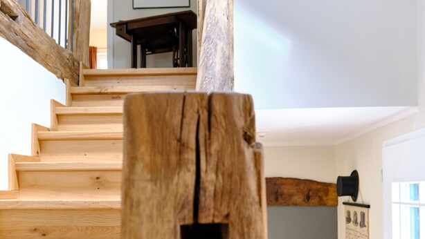 Un escalier en bois au design rustique, menant à une pièce dont le sol est recouvert d’un parquet à motifs en chevrons et qui contient une chaise en bois.