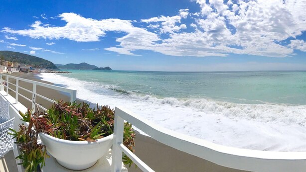 A seaside balcony with a white railing, a potted plant, and a view of the ocean and distant hills.