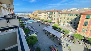 A courtyard with outdoor seating, surrounded by buildings with balconies and a view of the sea.