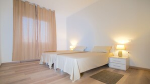 A bedroom with two beds, a nightstand with a lamp, a window with curtains, and a wooden floor.